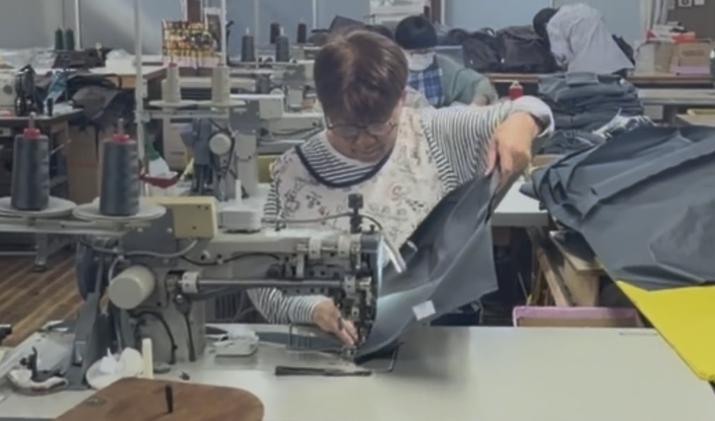 縫製工場内で、作業者が大型の工業用ミシンを使い、厚手の黒い生地を縫製している様子。周囲には糸や裁断済みの生地が並び、奥にも複数の作業台やスタッフが見える工場風景。